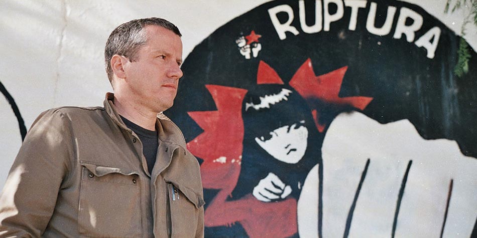 Presenter Carne Ross standing in front of an anarchist mural