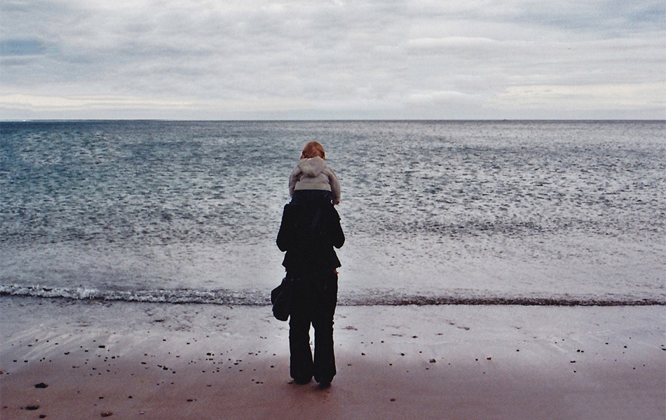 The back of a woman with her son on her shoulders looking out at sea.