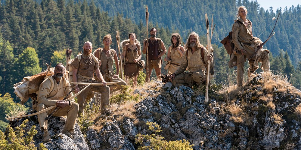 Male and female Stone Age experts, dressed as their ancestors, carrying spears and bows on a mountain top in forested wilderness