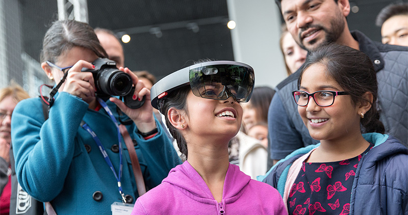 A smiling girl wearing Virtual Reality Headsets at the Great Exhibition Road Festival 2019 surrounded by spectators.