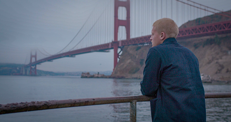 A young man with short red hair, wearing a blue jacket, in semi profile from behind with a suspension bridge in the background.