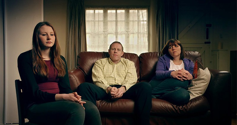 Daughter sits in chair in foreground while parents sit on sofa in the background