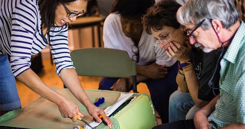 Female artist demonstrates how to upholster a chair to three onlookers