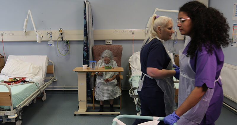 Hospital staff working on a ward while a patient sits in the background